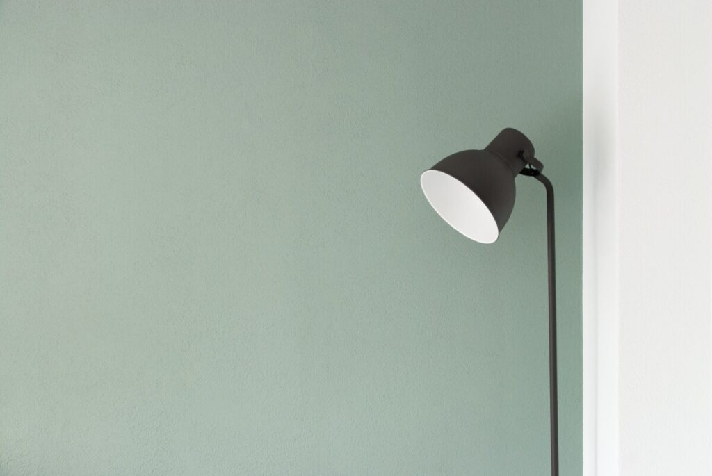 A minimalist designed room featuring a mint green wall with a simple, elegant black floor lamp. The lamp's design is modern, with a curved neck and a sleek, round shade, providing a contrast against the plain, textured background. The composition conveys a sense of simplicity and functional design.